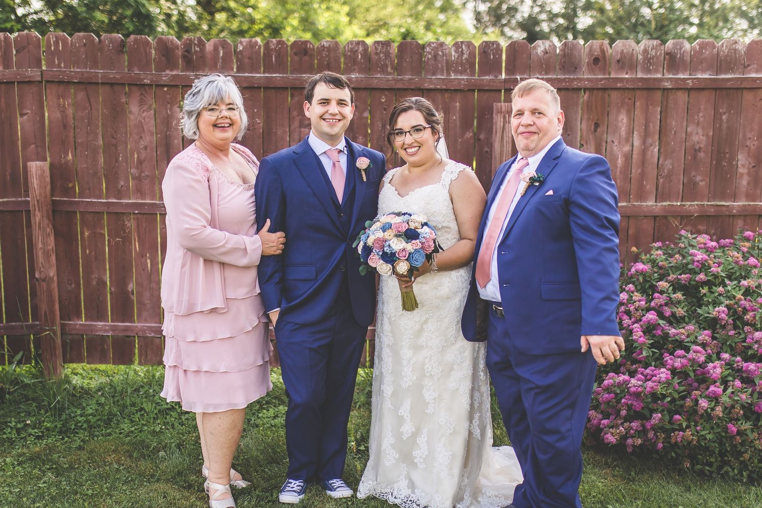 Wedding group portrait against a wooden fence with pink flowers blooming in the background.