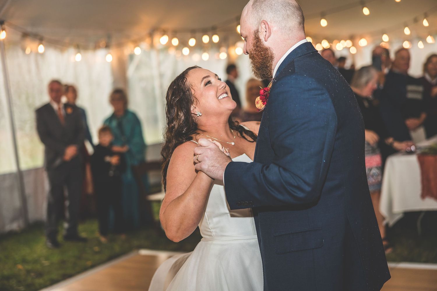 Couple shares a romantic dance at a wedding reception under twinkling string lights in an elegant tent setting.