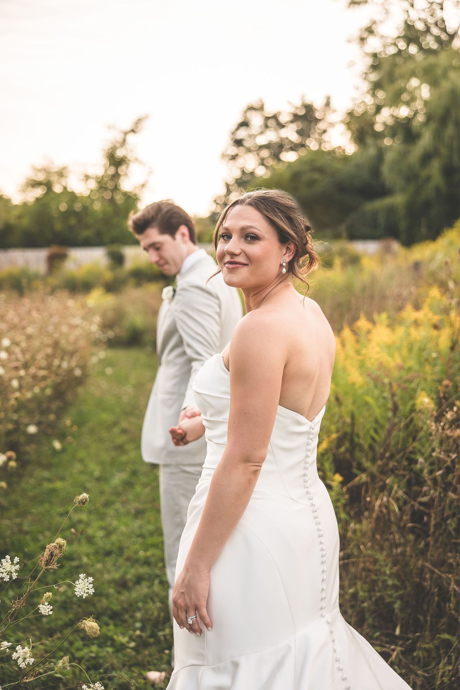 Wedding couple walks through a wildflower meadow near a rustic red barn at sunset.