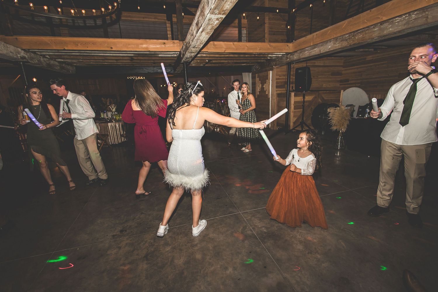 Wedding reception dance party in rustic venue with string lights and wooden beams.