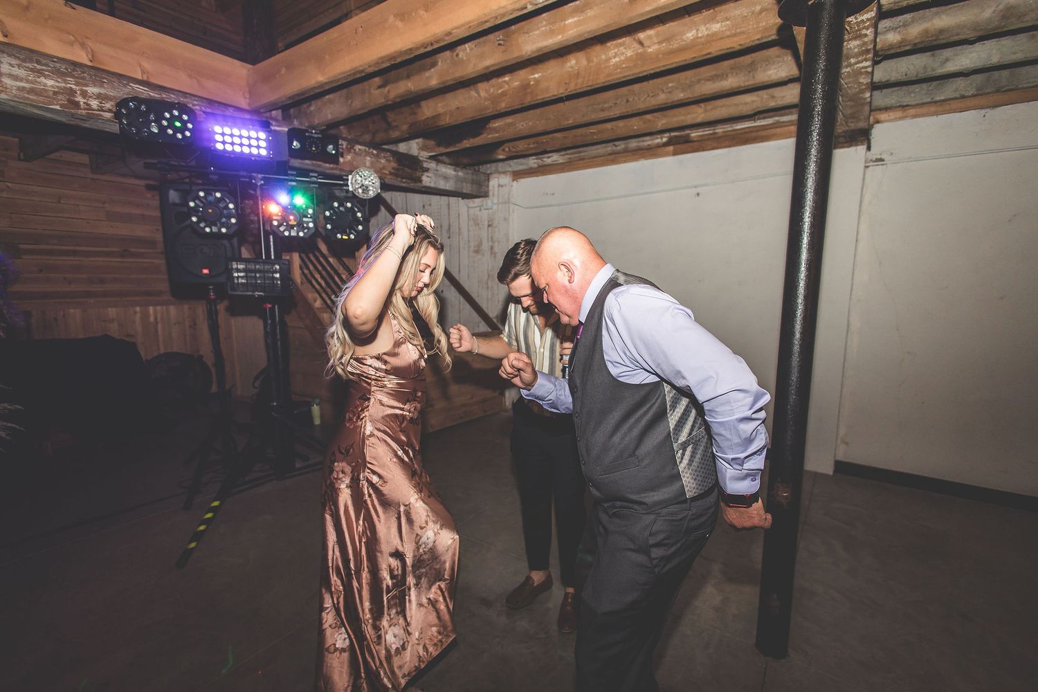 Couples dancing at a wedding reception in a rustic venue with exposed beams and mood lighting.