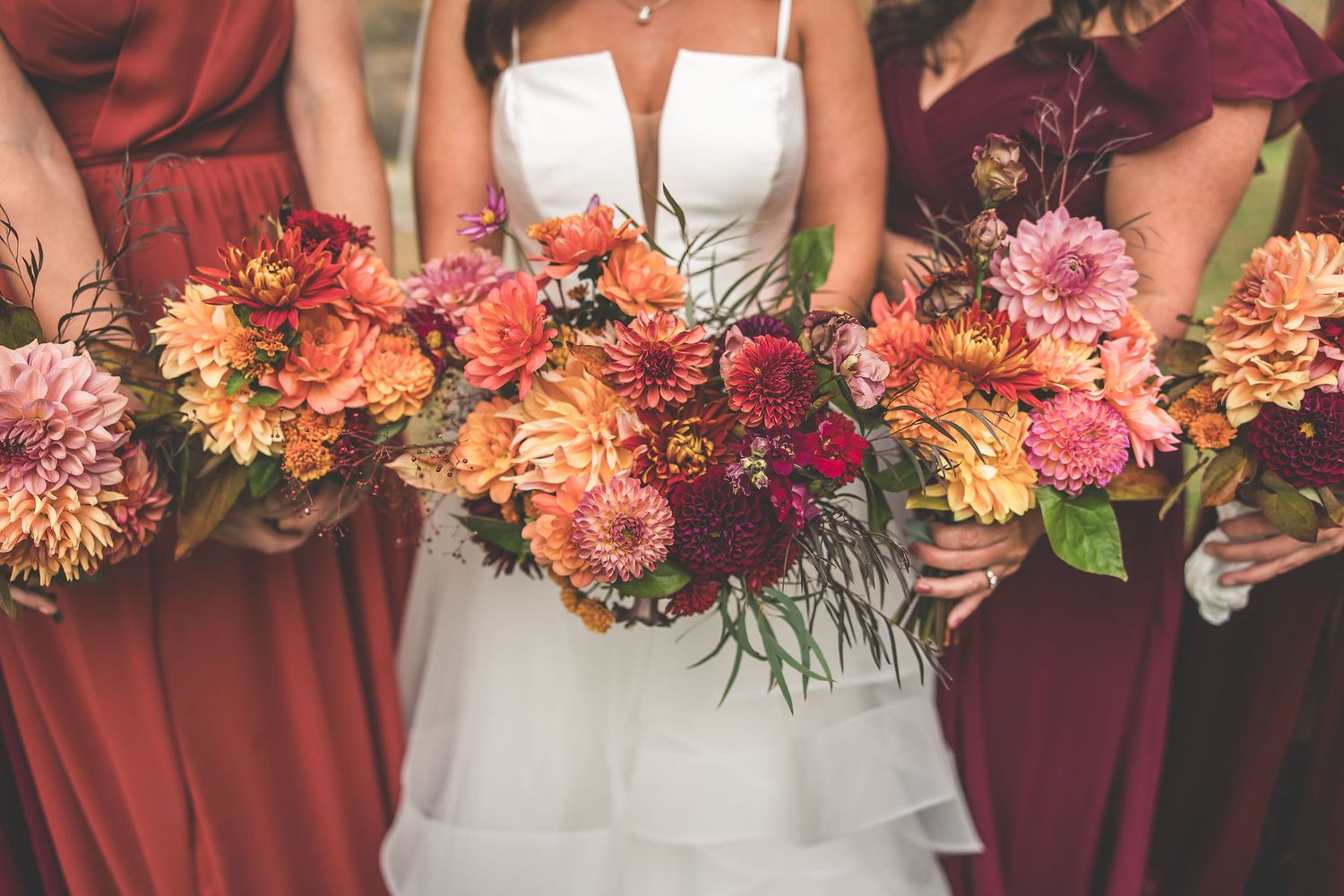 Vibrant coral and burgundy floral bouquets held by wedding party members in autumn colors.