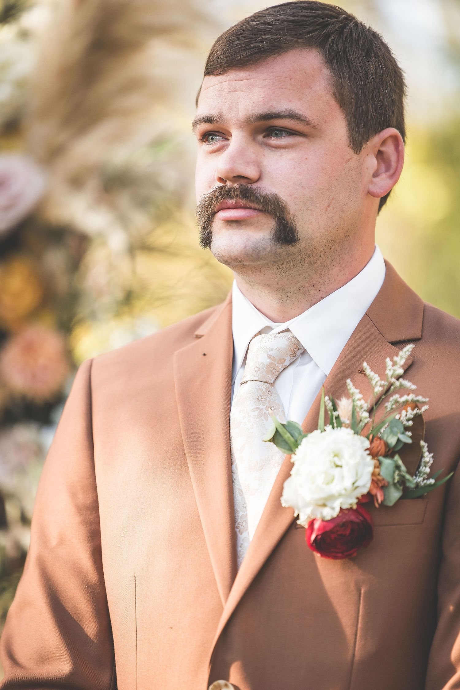 Groomsmen wearing brown suits with white boutonnières stand in a row outdoors.
