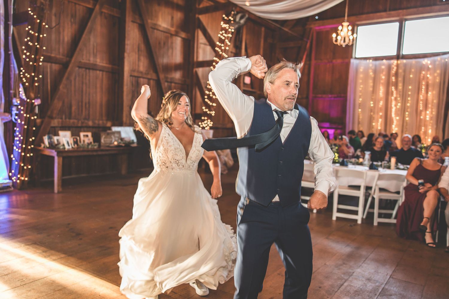 Wedding couple and guests dance together on a rustic wooden dance floor in a barn venue with string lights.