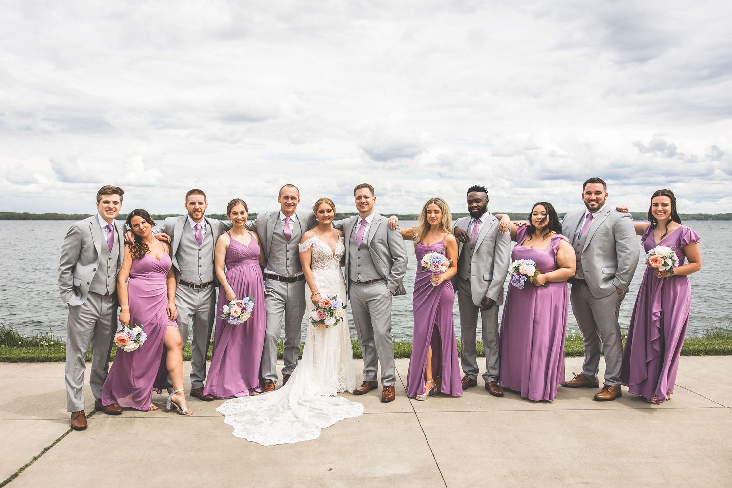 Wedding party in grey suits and mauve dresses posed by waterfront.