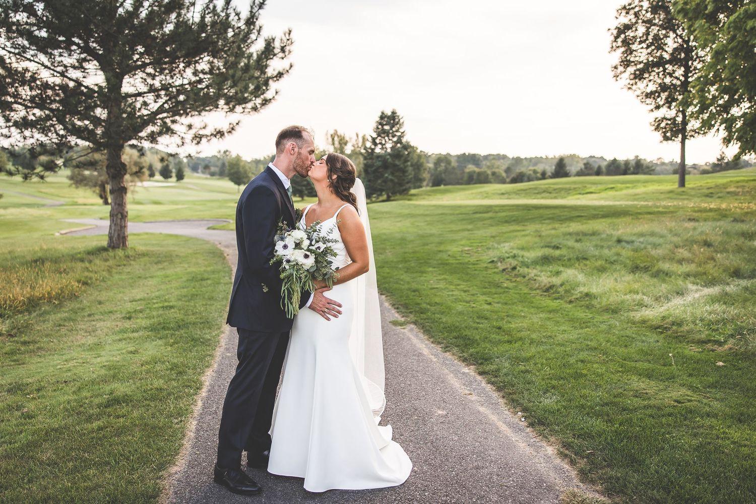 Wedding couple walks along winding path on golf course during sunset photo session.