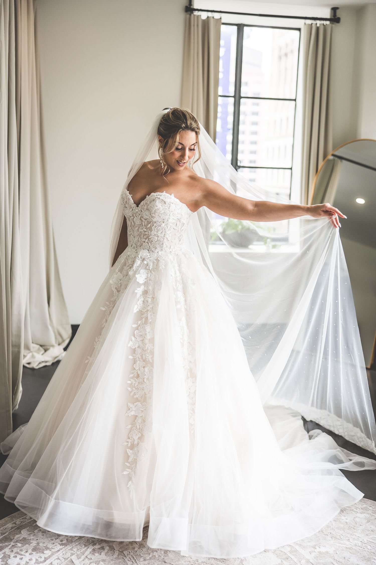 Elegant bride adjusts her flowing white wedding gown in front of window.