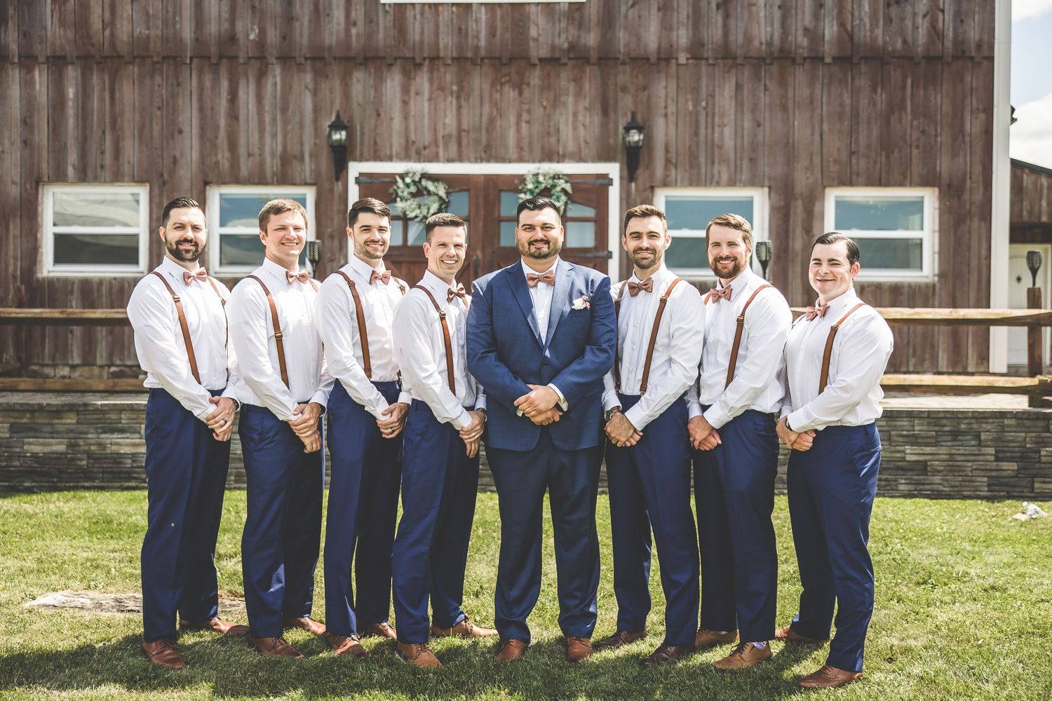 Groomsmen in navy suits and suspenders posing playfully outside rustic barn.