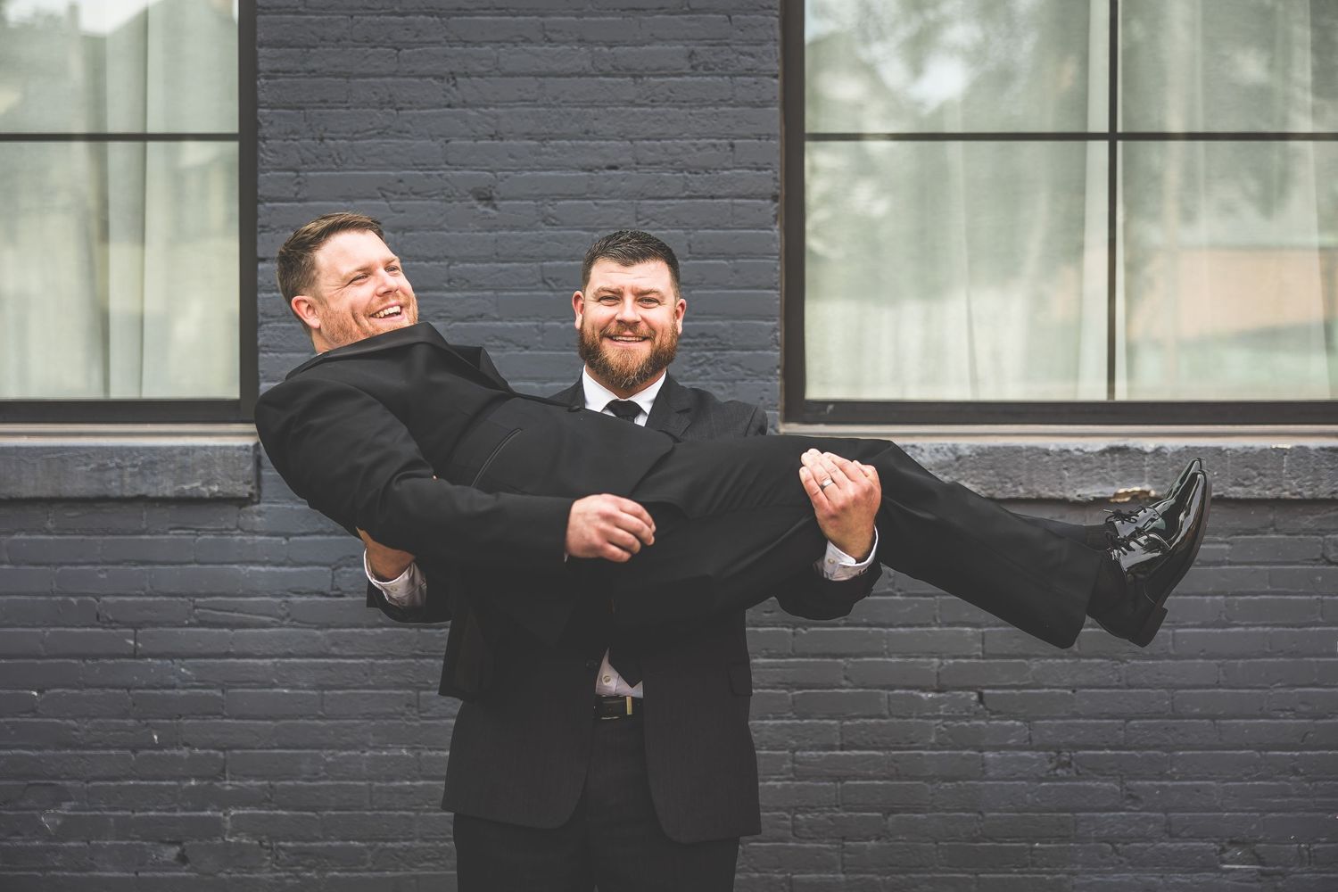 Wedding party members playfully lift and carry each other in front of brick wall.