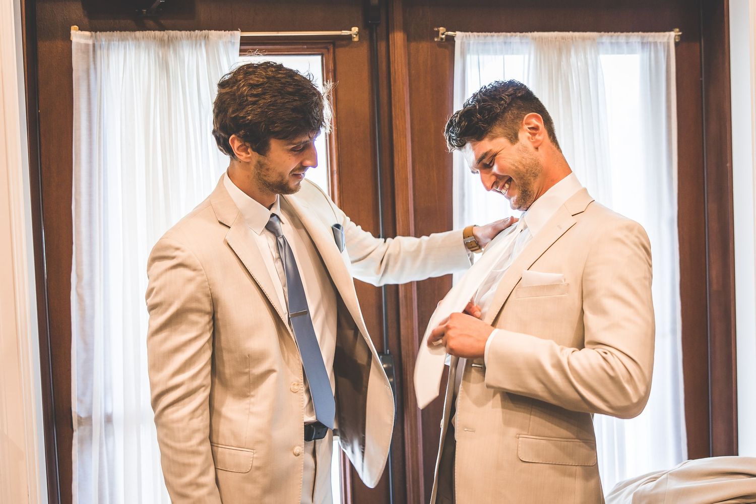 Men in beige suits adjusting their attire in wooden-framed mirrors.