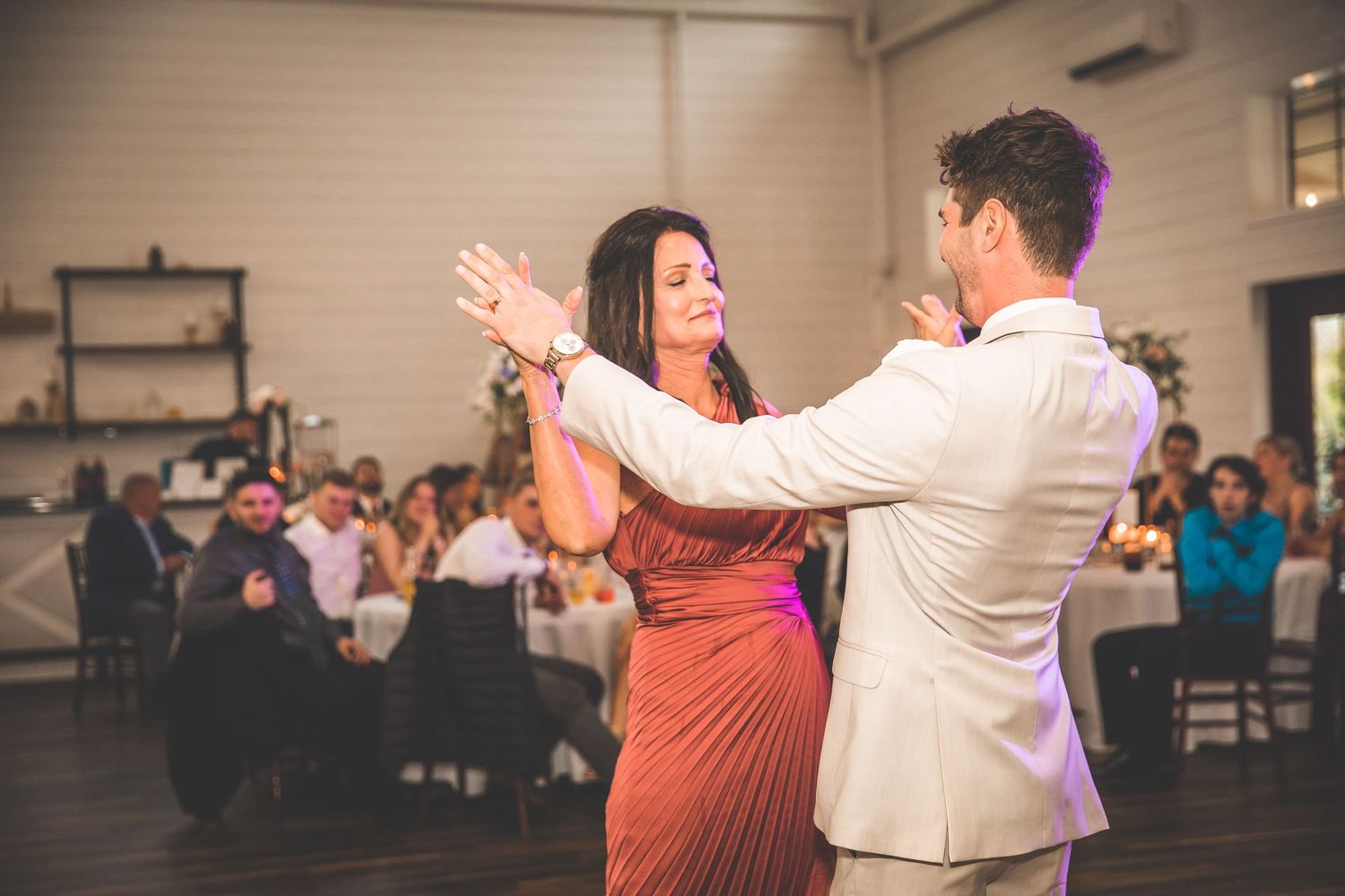 A couple dances together at a wedding reception, with the woman wearing a coral dress and man in a light suit.