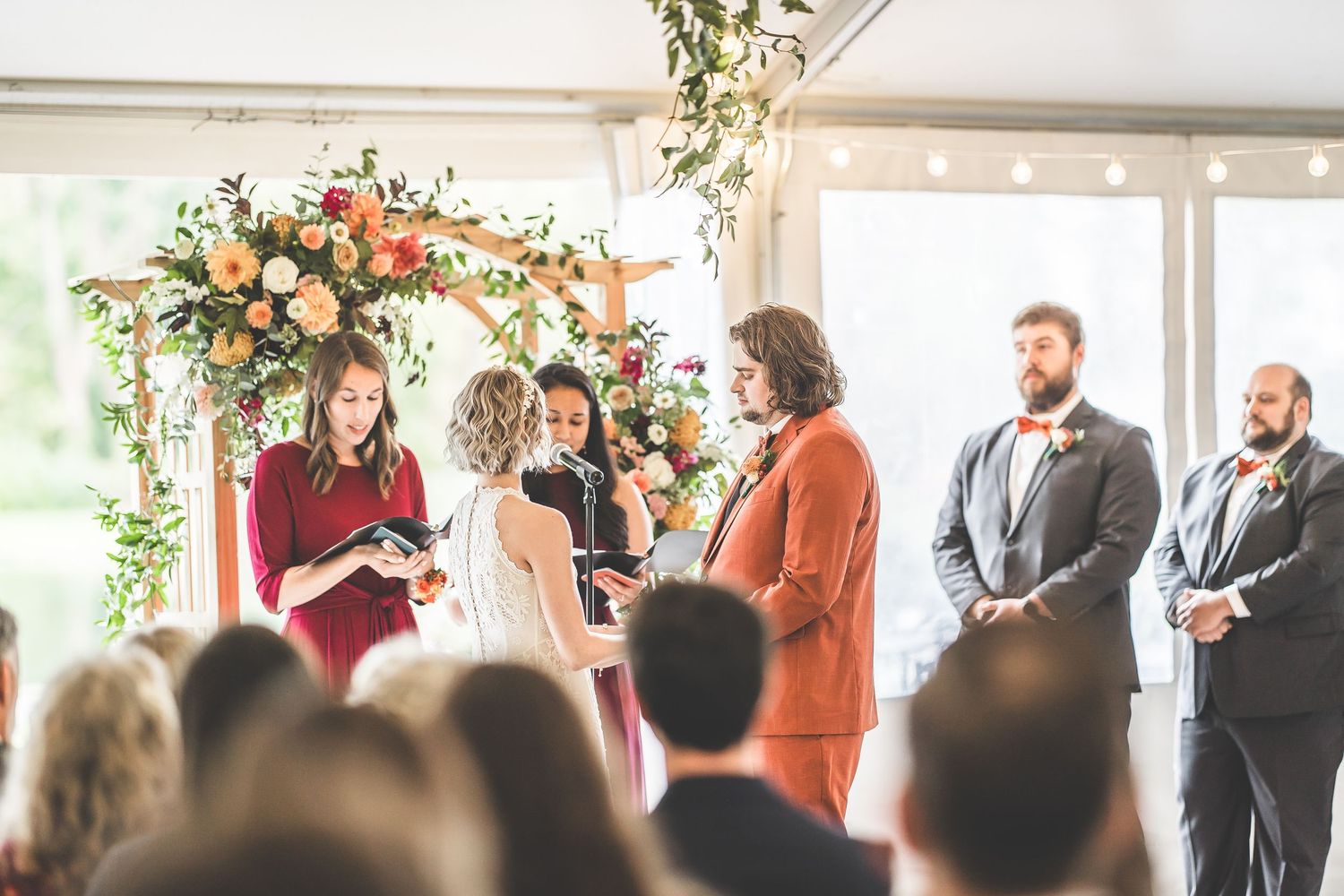 Wedding guests observe ceremony proceedings in bright indoor venue with floral decorations.