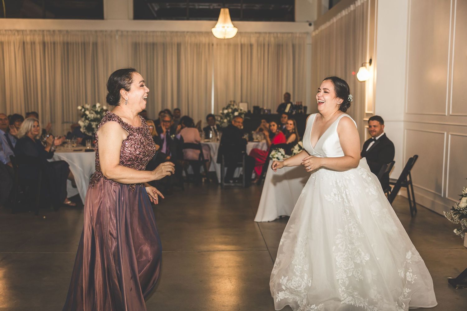 Wedding guests dance and celebrate on an elegant ballroom dance floor with warm lighting and white drapes.