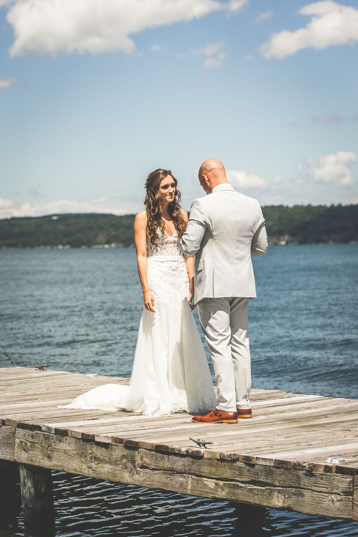 A bride in a flowing white dress stands with her groom on a wooden dock by the water.
