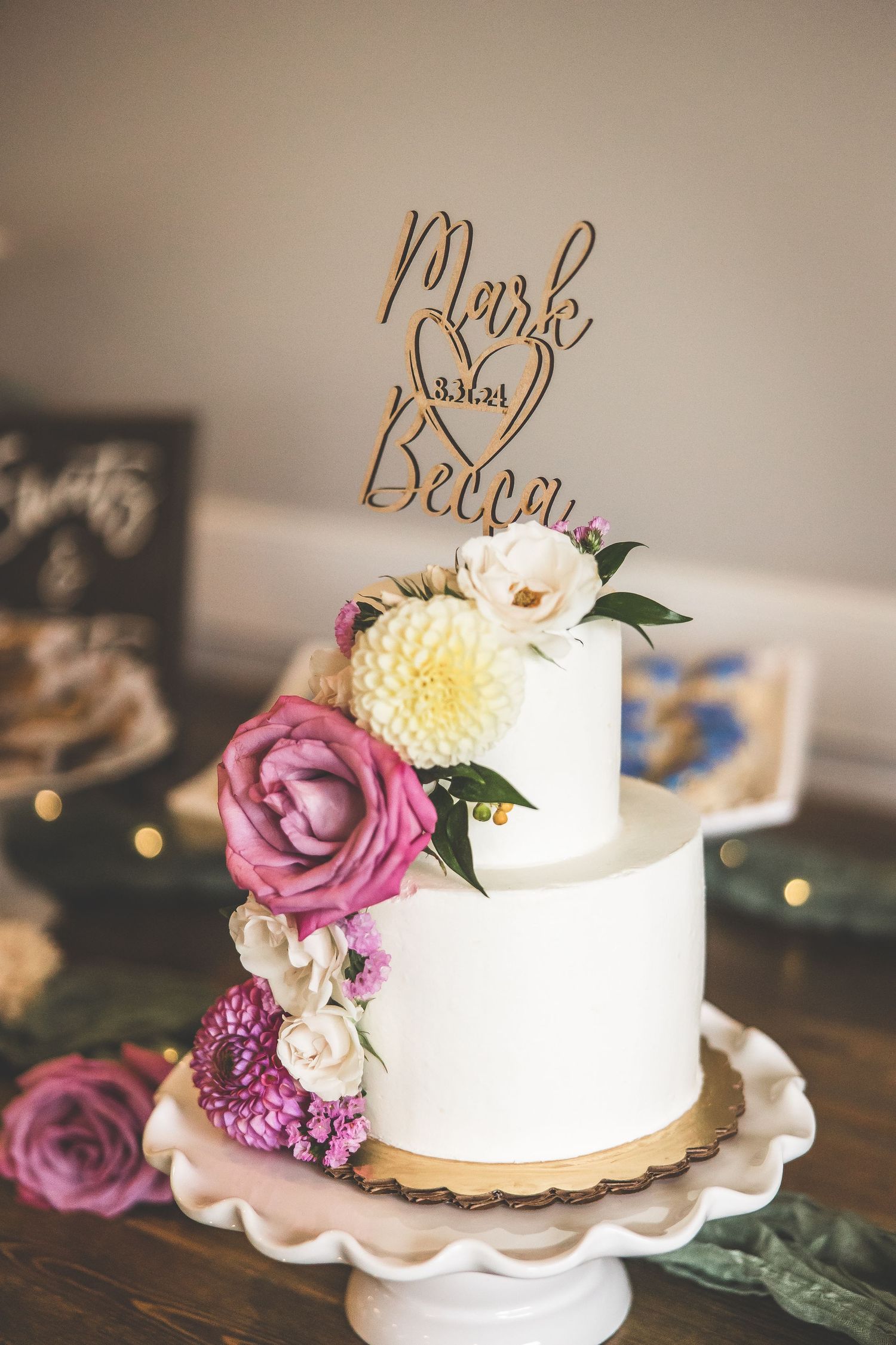 White wedding cake decorated with pink roses and gold script topper on a cake stand.