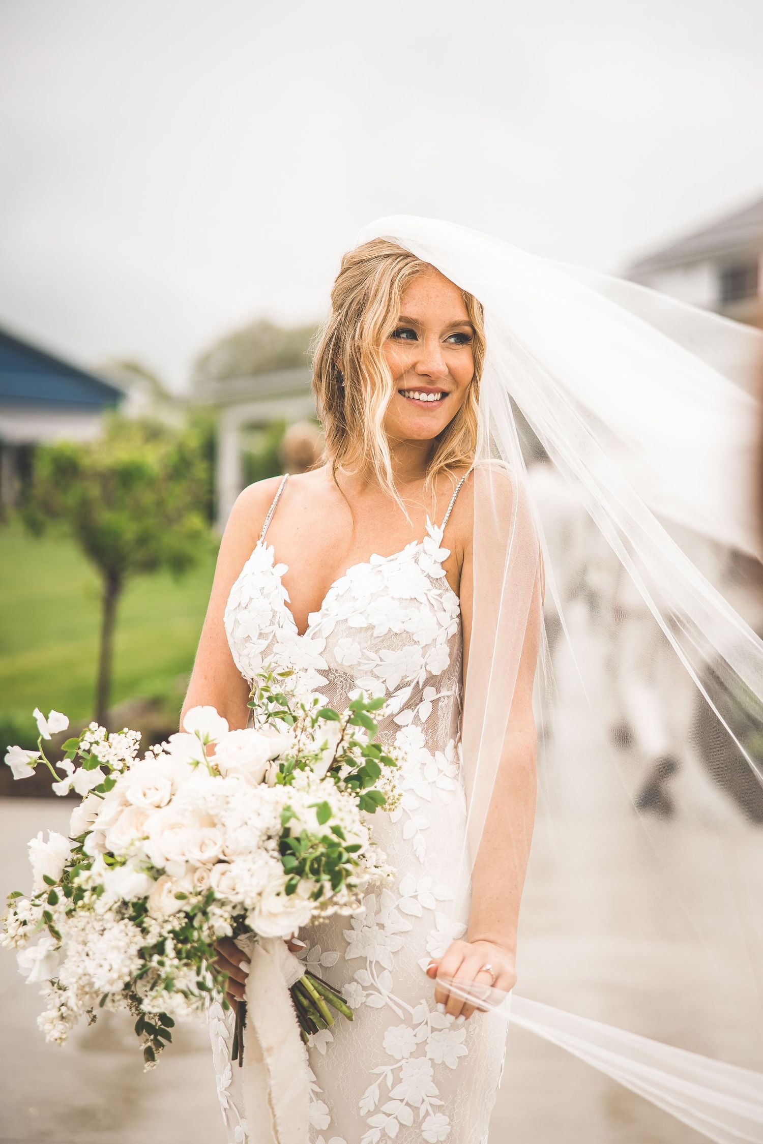 Bride in lace dress holds white bouquet while veil flows in the breeze.