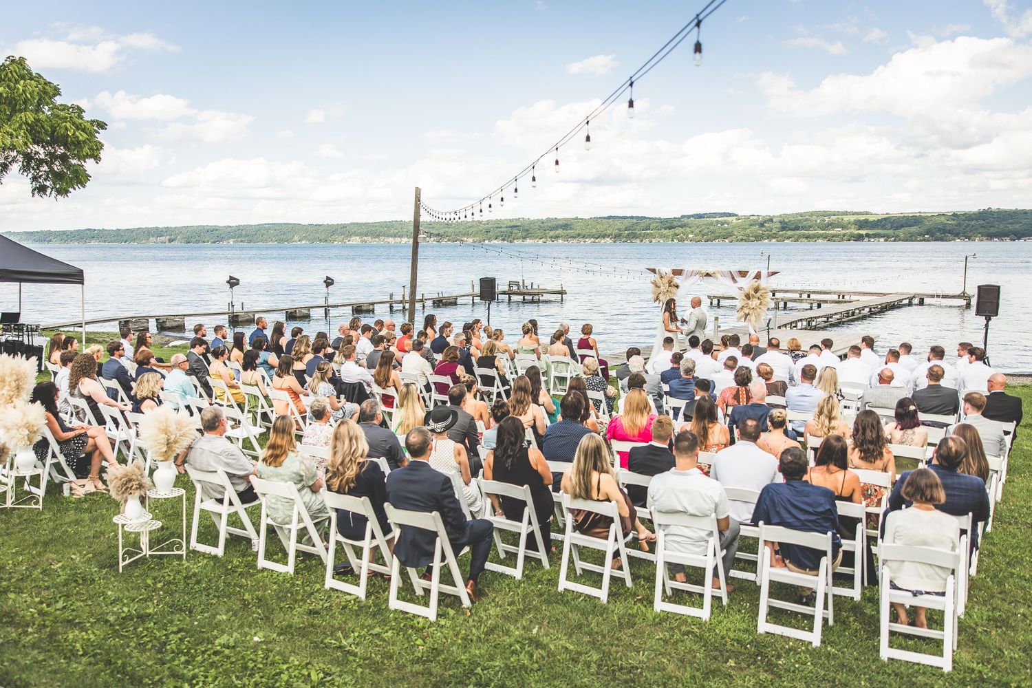 Panoramic view of lakeside wedding ceremony setup with white chairs and string lights overhead.