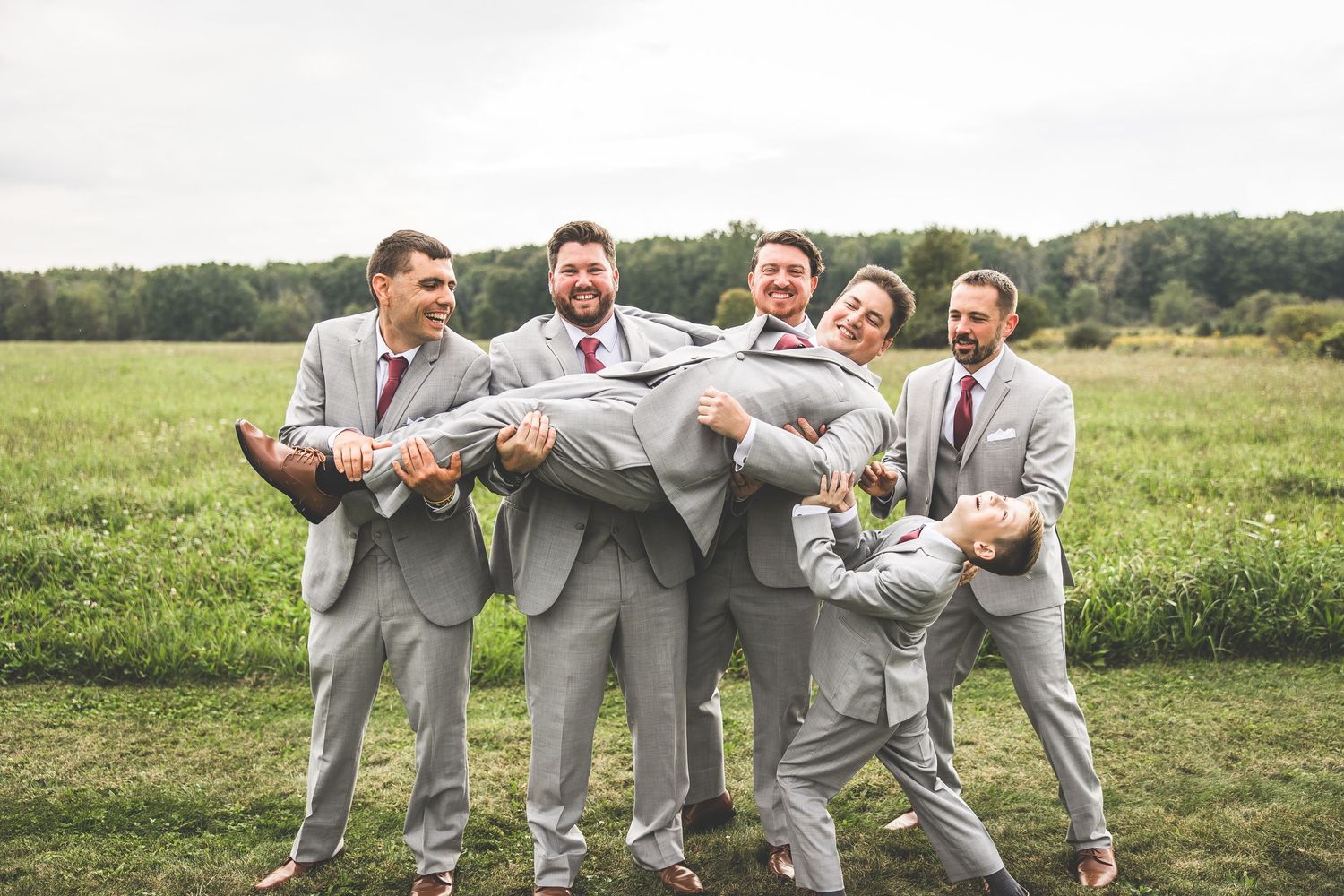 Groomsmen in matching gray suits playfully carry the groom in a field.