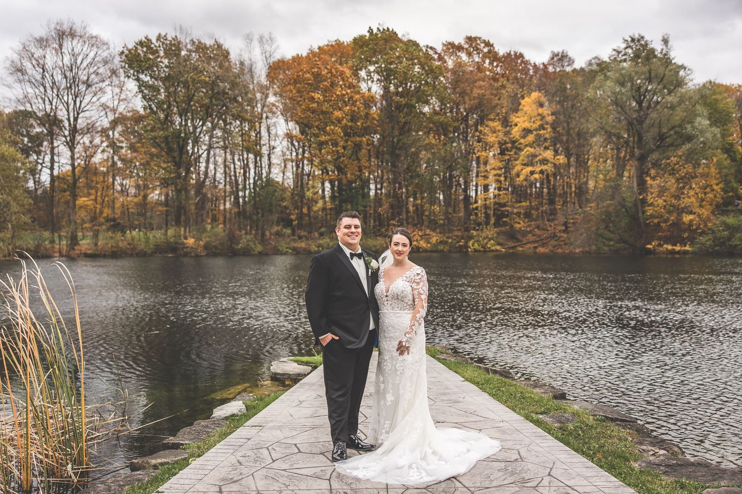 Wedding couple poses on stone path by lake with fall foliage reflecting in water.