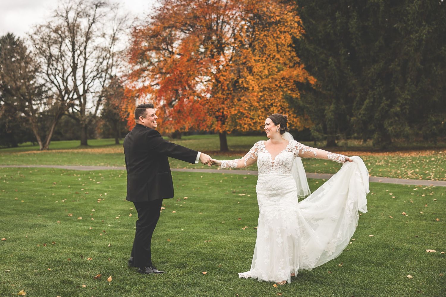 Wedding couple dances and twirls on green grass with autumn trees in the background.