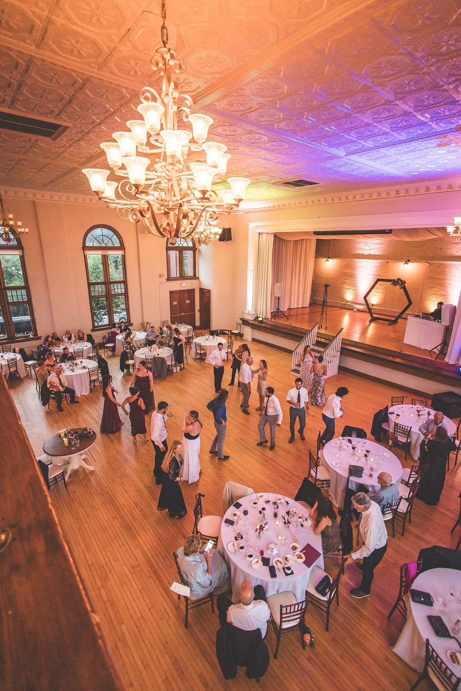 Wedding reception venue with elegant chandeliers and hardwood floors filled with guests mingling at decorated tables.