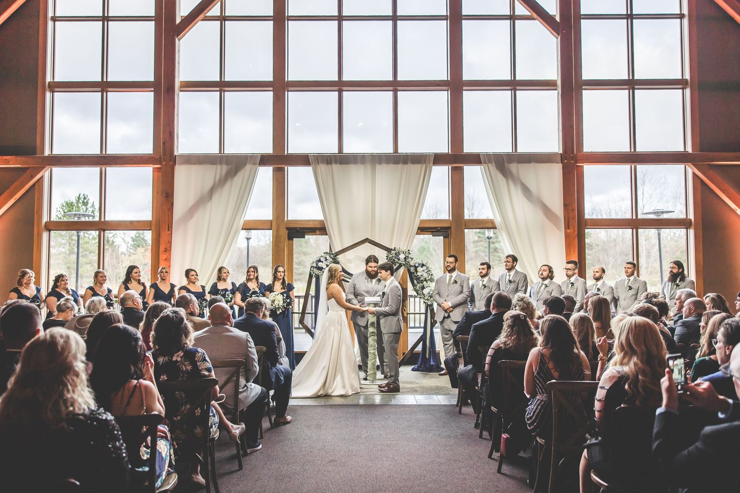 Panoramic view of wedding ceremony in a sunlit venue with floor-to-ceiling windows and wooden architecture.