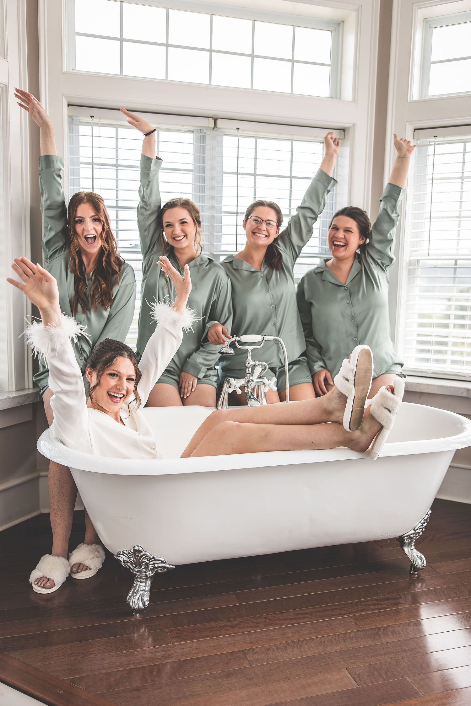 A group in matching sage green sweatshirts poses playfully around a white clawfoot bathtub in a bright room.