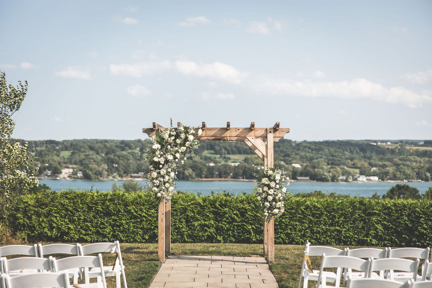 Outdoor wedding ceremony setup with wooden arch and white chairs overlooking a serene lake view.