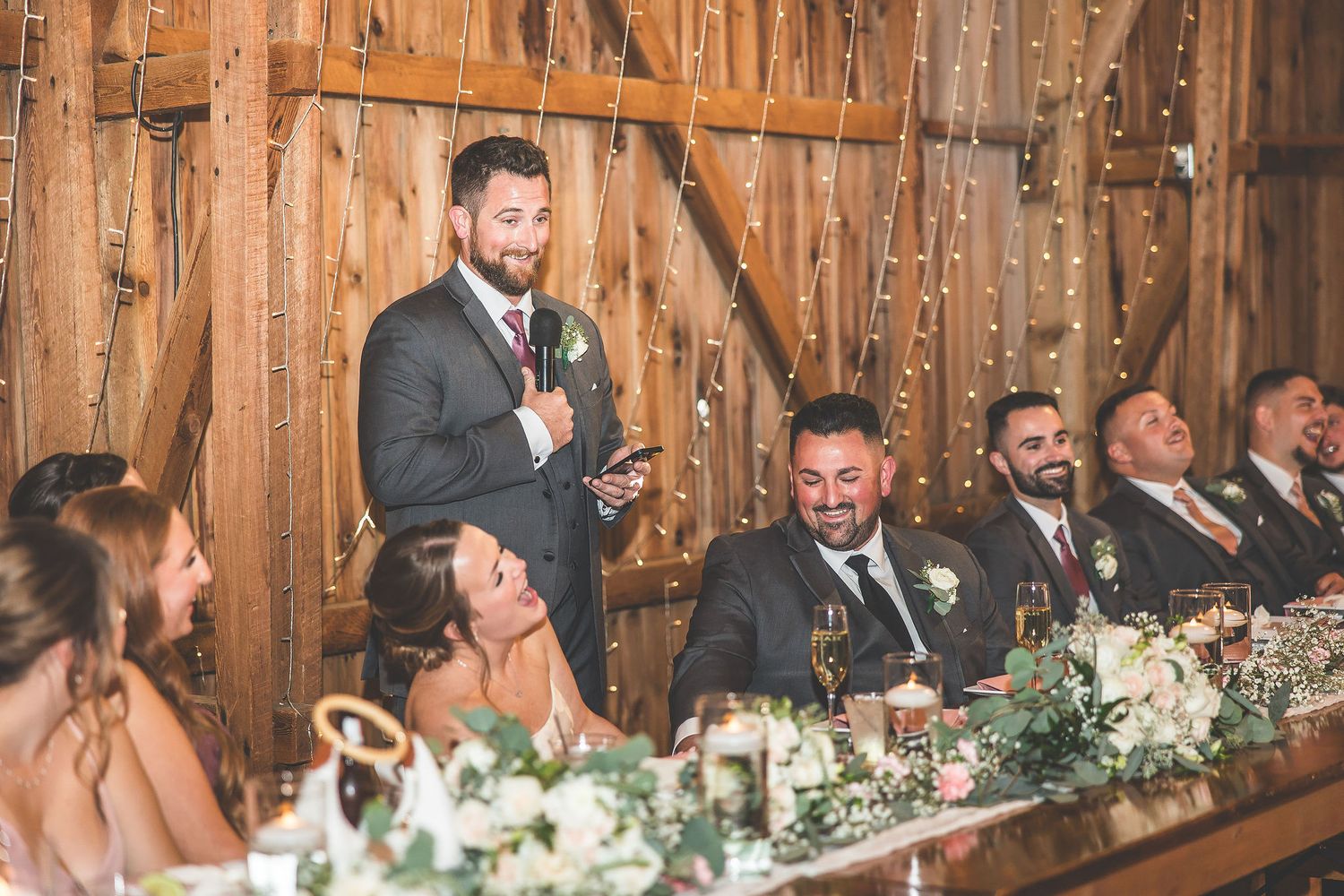 Wedding party seated at decorated head table in rustic barn venue with string lights.
