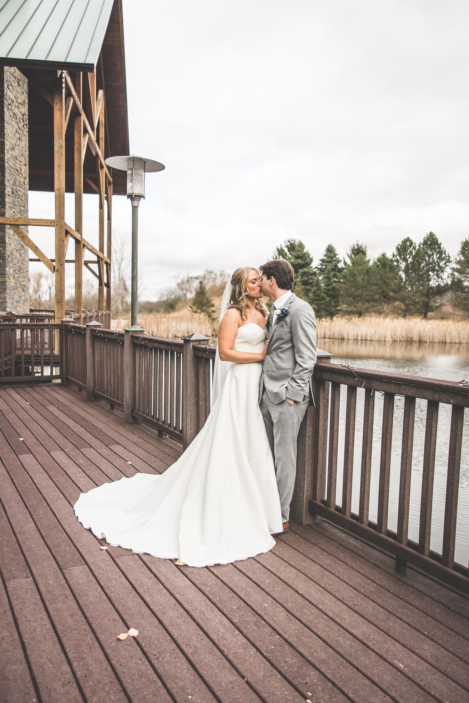 Wedding couple shares intimate moment on rustic wooden deck overlooking water during autumn day.