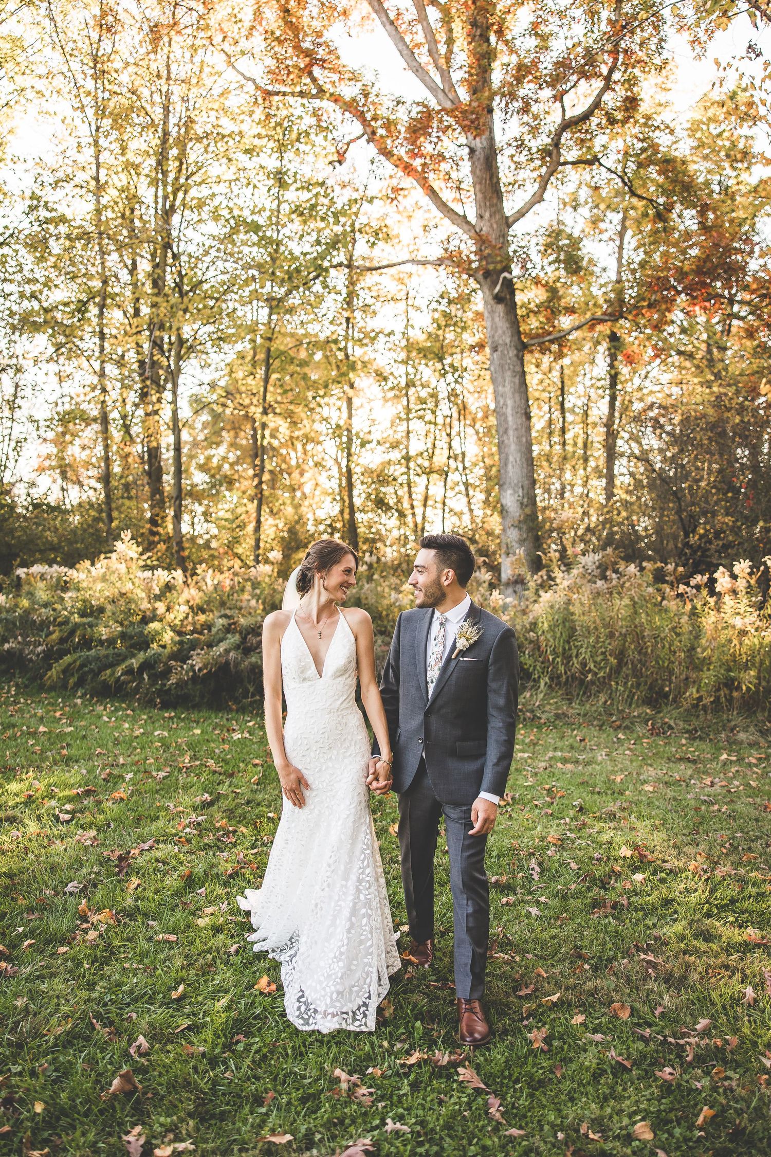 Elegant couple in wedding attire standing together in autumn woodland with sunlit trees.