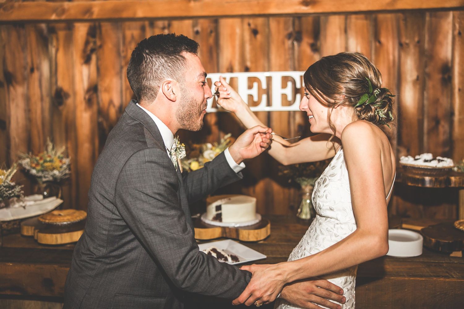 Newlyweds enjoying cake and desserts at rustic wooden reception table.