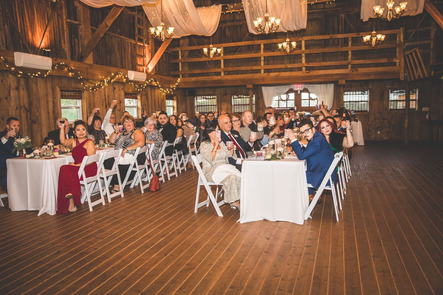 Wedding guests seated at tables in a spacious barn venue decorated with hanging lights and white linens.