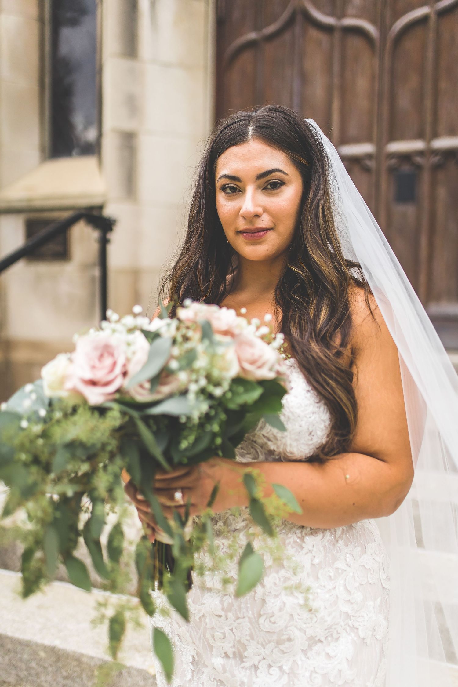 Bride holds romantic bouquet of pink roses and greenery while wearing a lace wedding gown and veil.