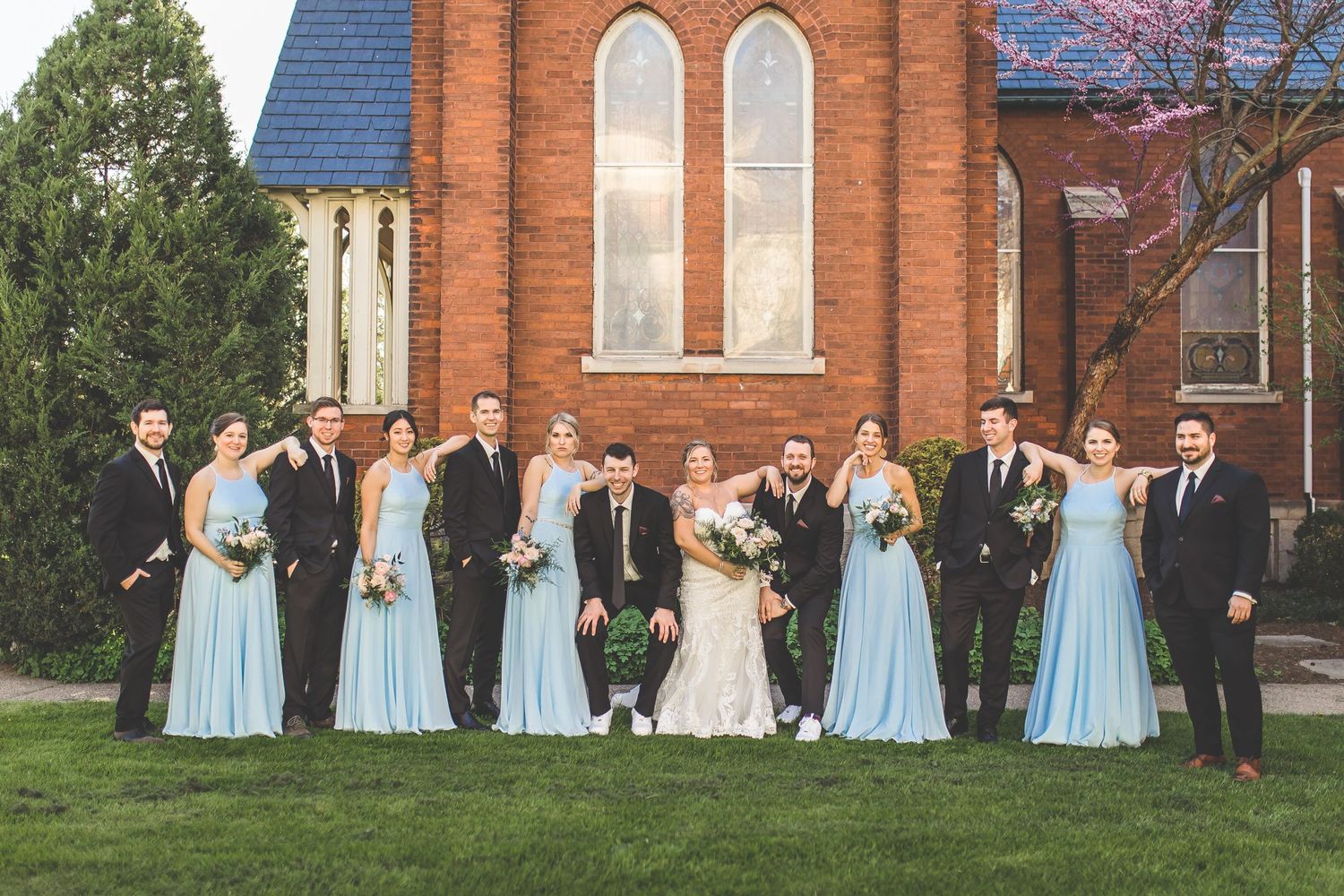 Wedding party poses on lawn with bridesmaids in light blue dresses and groomsmen in dark suits.
