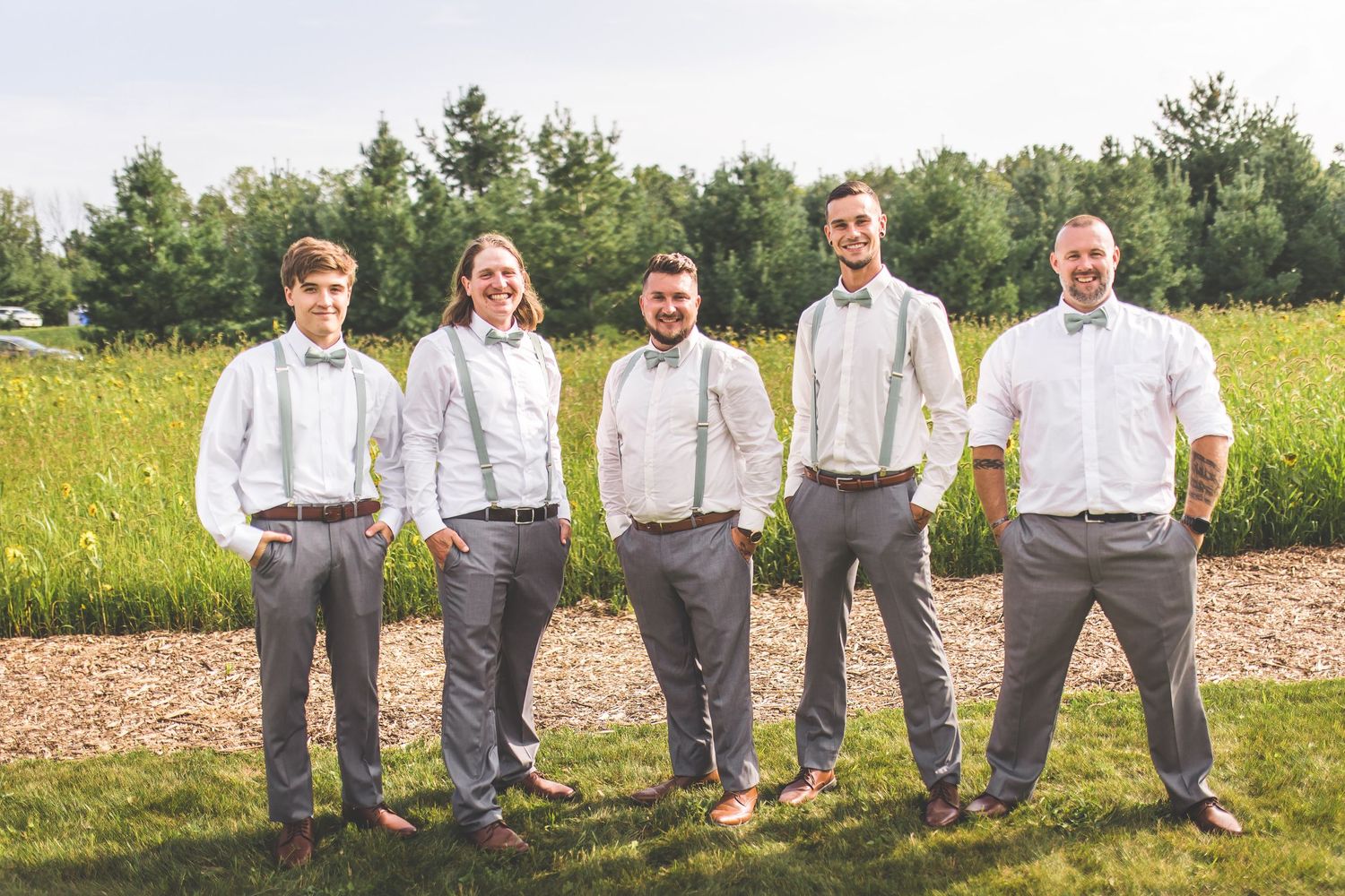 Wedding party members in grey pants and suspenders stand in a line outdoors.