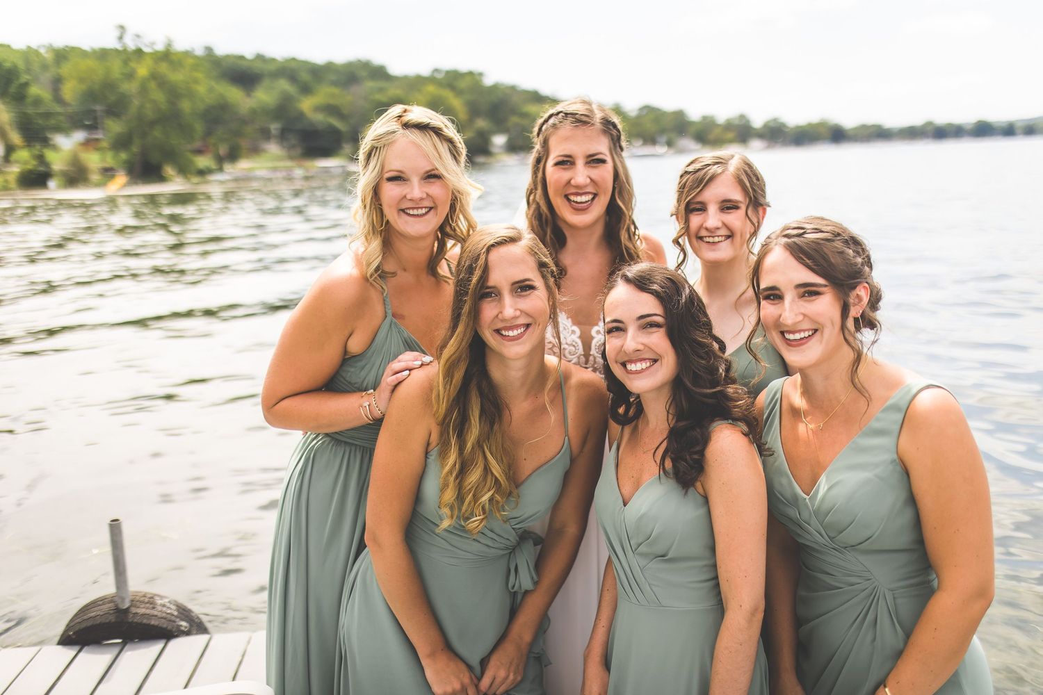 Bridesmaids in matching sage dresses smile together by lakeside on sunny day.