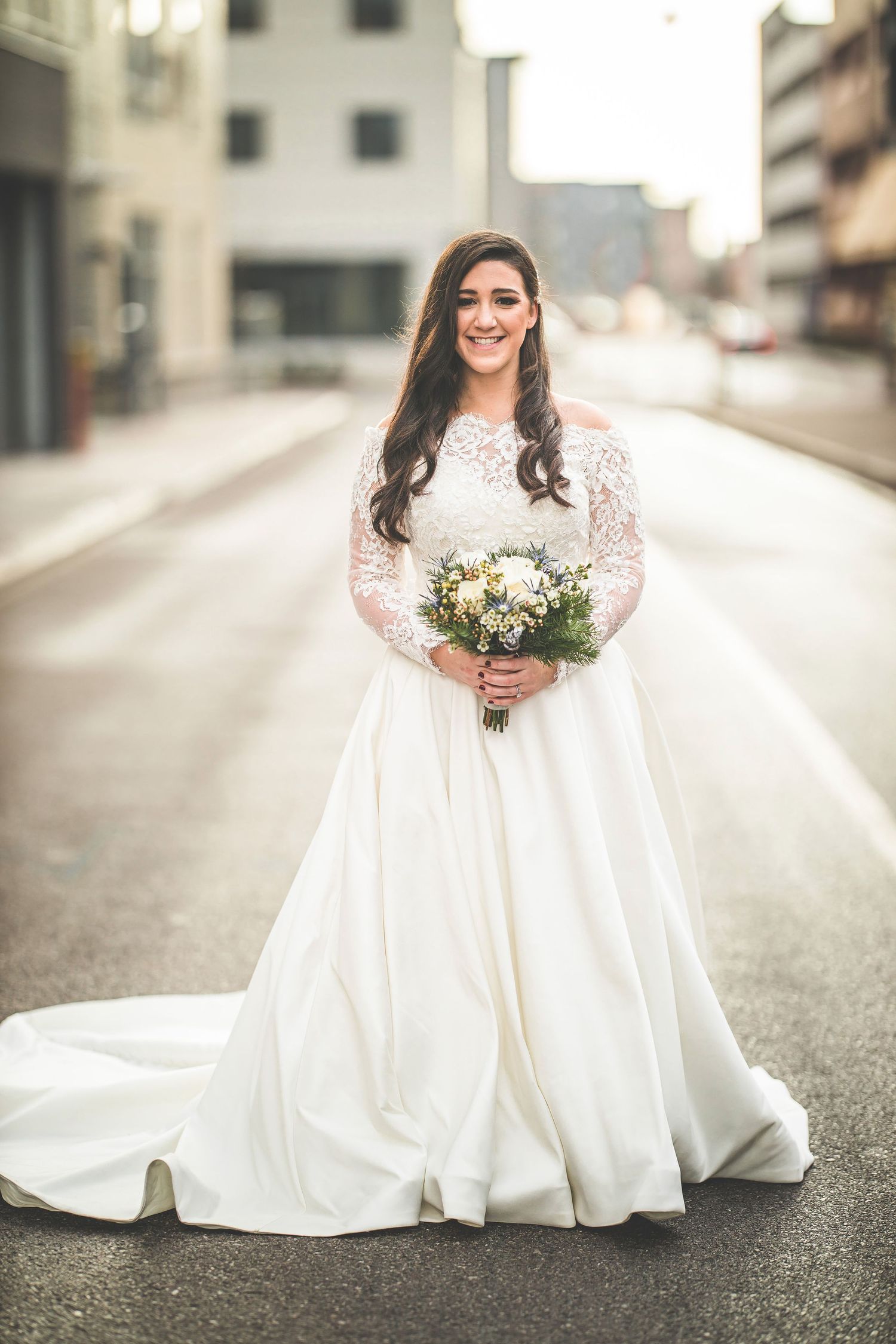 Bride in flowing white dress holding bouquet against modern building backdrop.