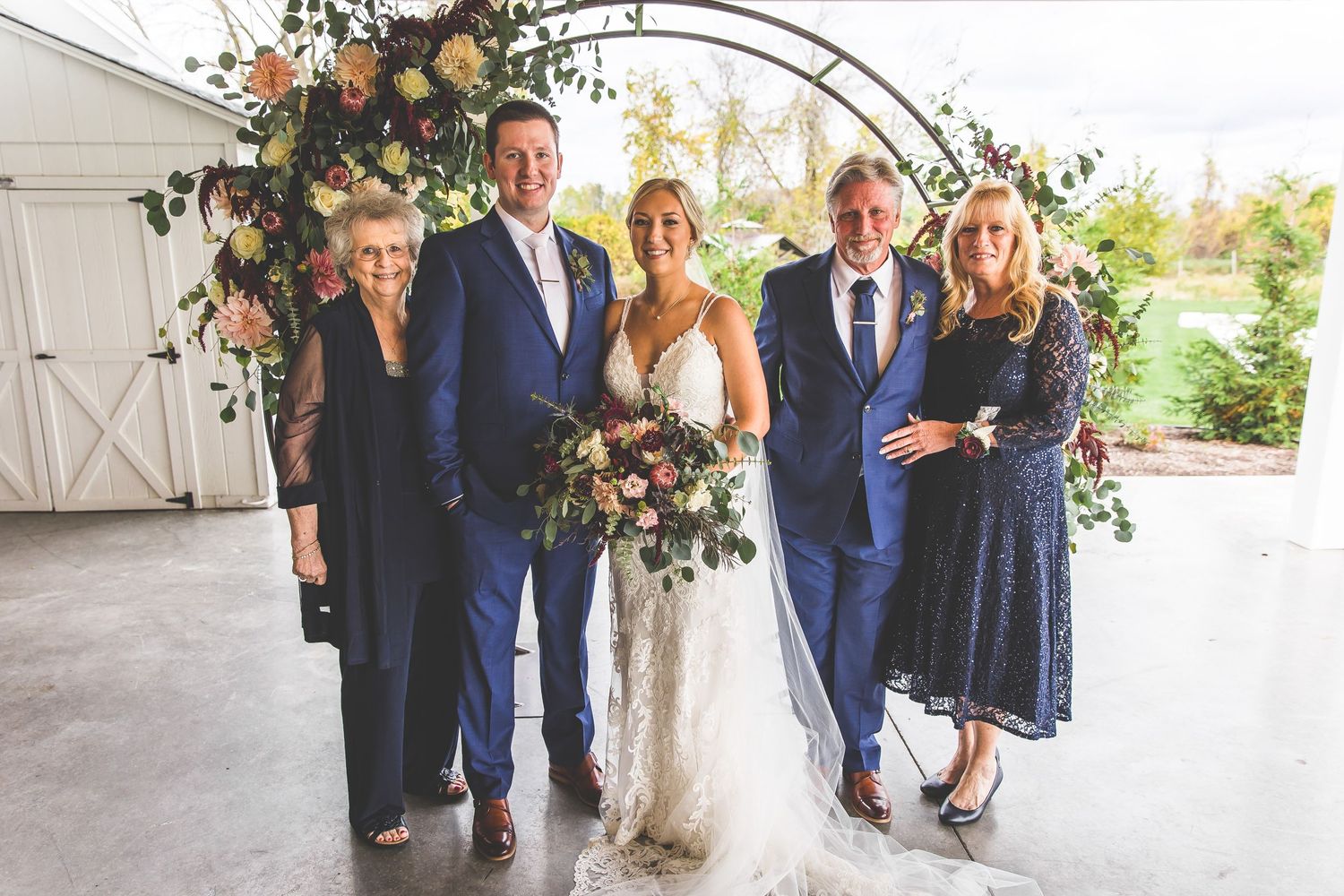 Wedding party gathers for formal photos under decorated floral archway.