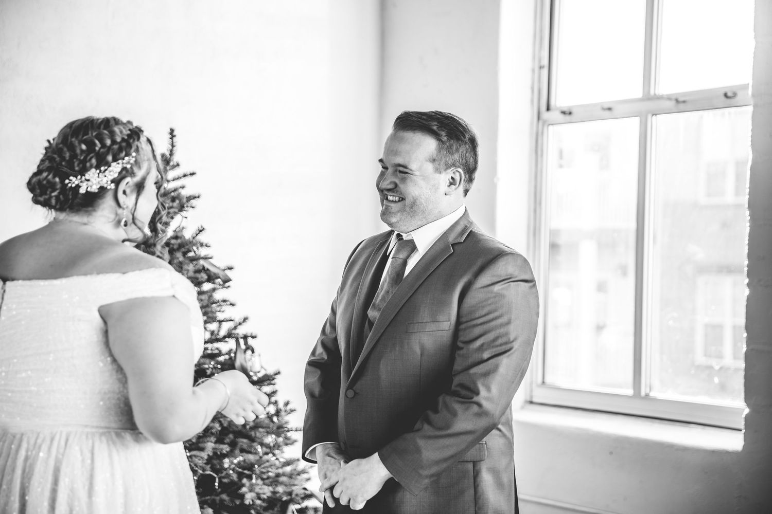 Black and white photo of wedding couple sharing first look moment by window with Christmas tree.