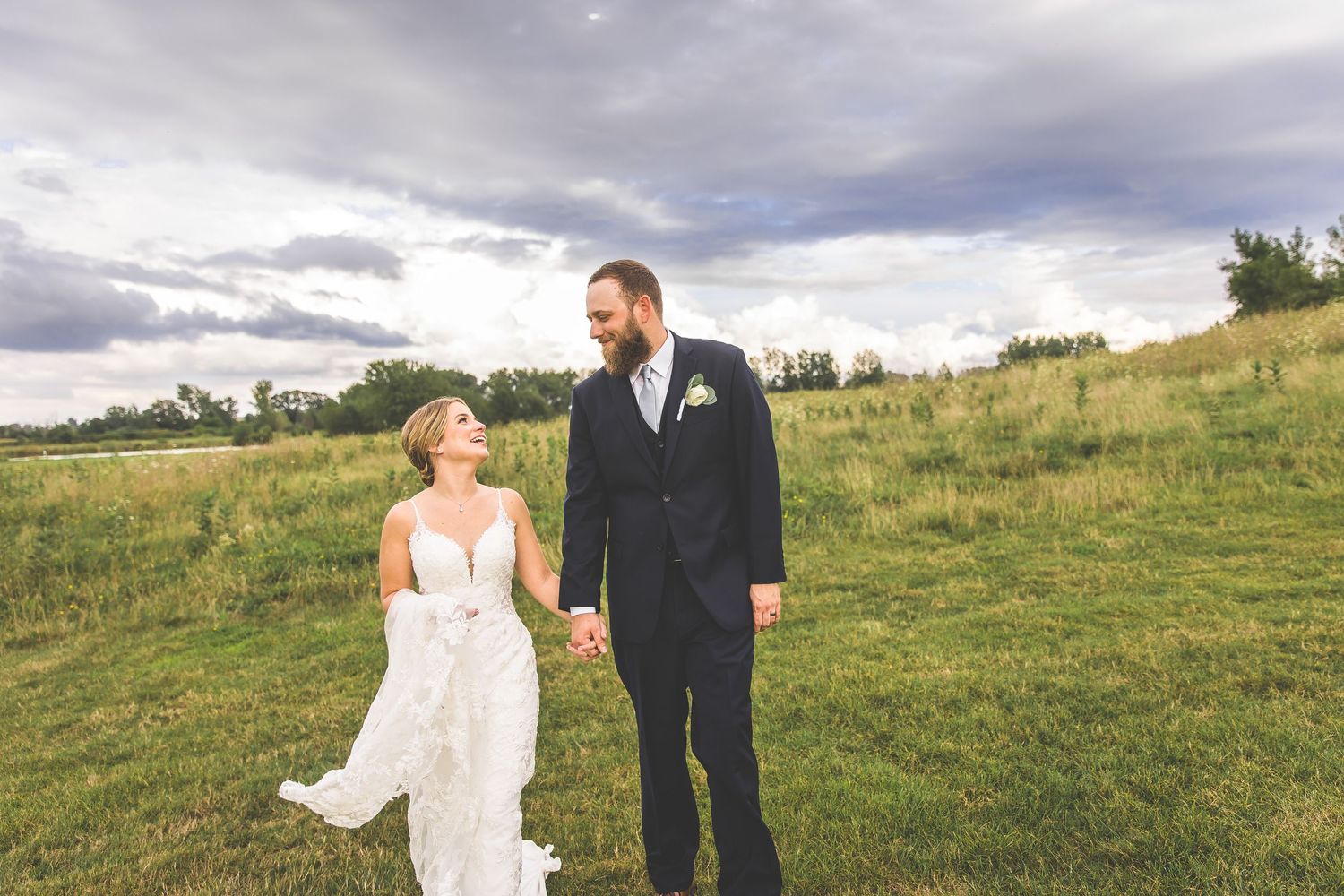Wedding couple walks hand in hand through a scenic meadow under cloudy skies.