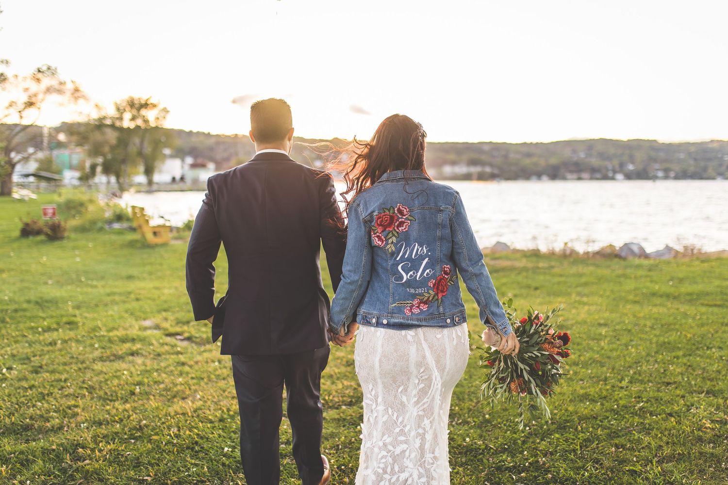 Bride in lace dress and denim jacket walks with groom along coastal path.