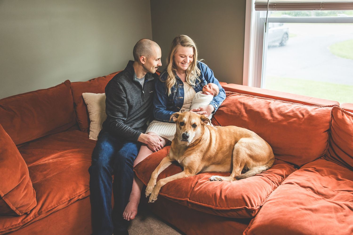 Couple relaxing on red couch with their brown dog while looking at mobile device.