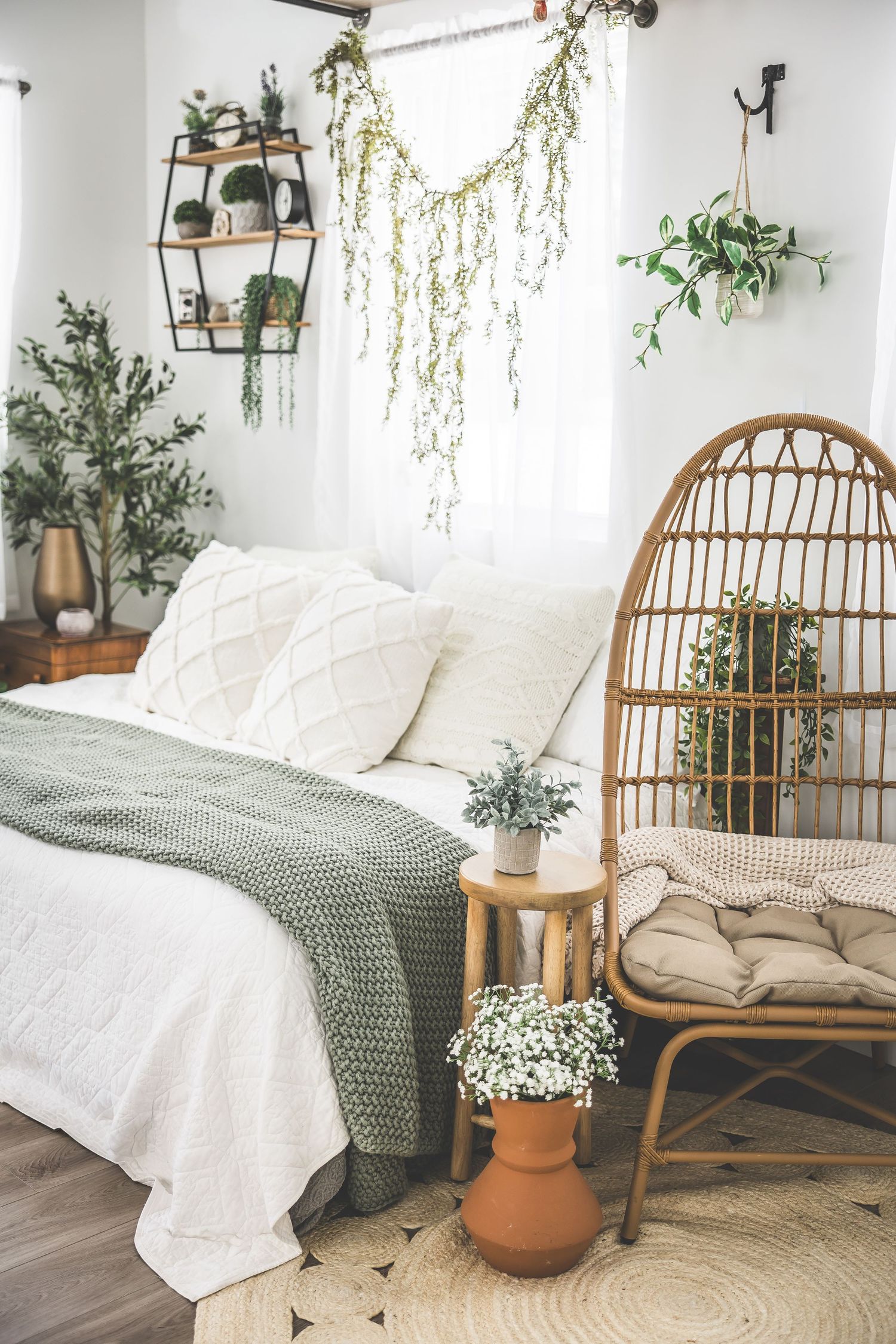 Cozy boho bedroom corner with rattan daybed, white bedding, green throw blanket, and hanging plants against white walls.
