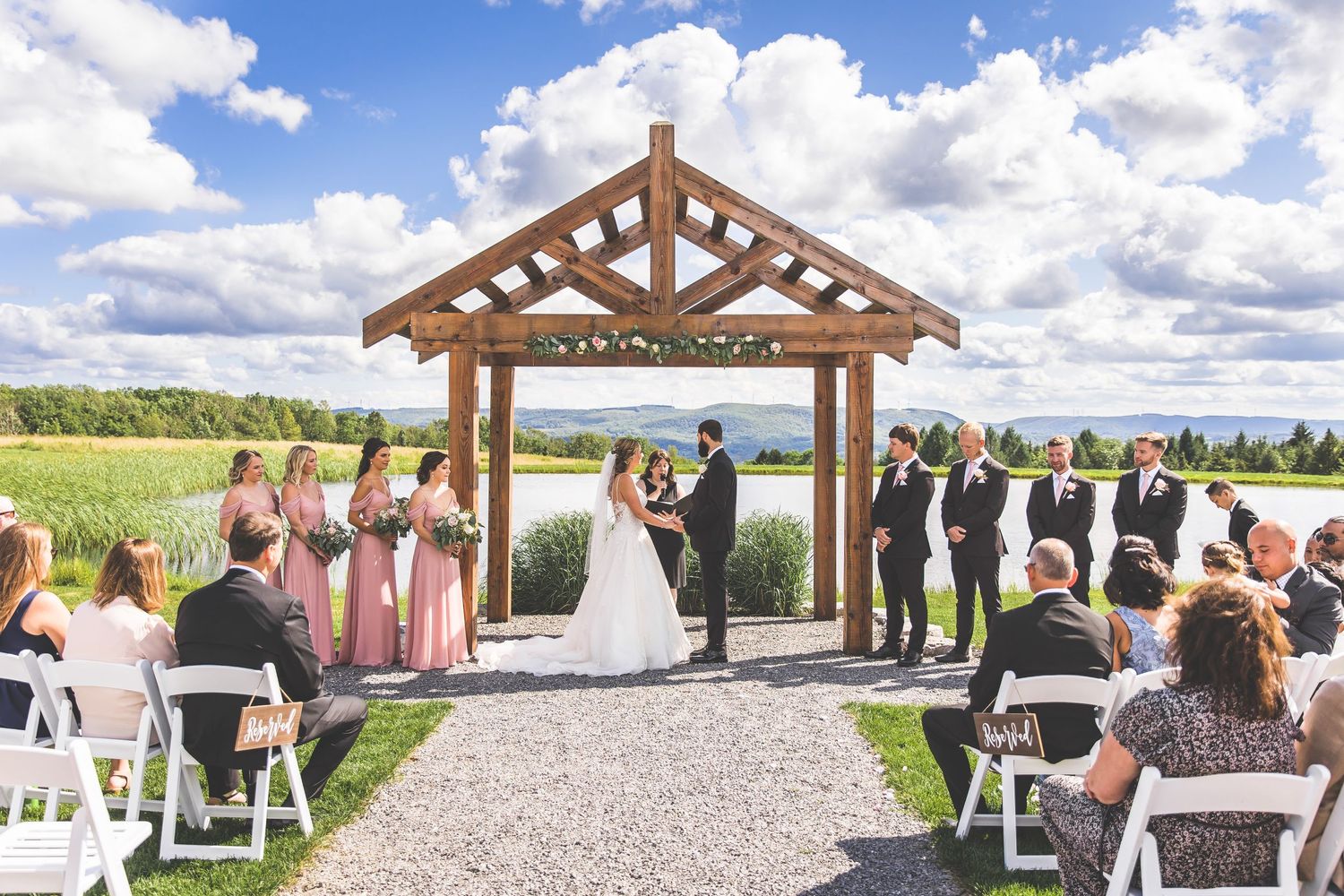 An outdoor wedding ceremony under a wooden pergola with guests seated on white chairs.
