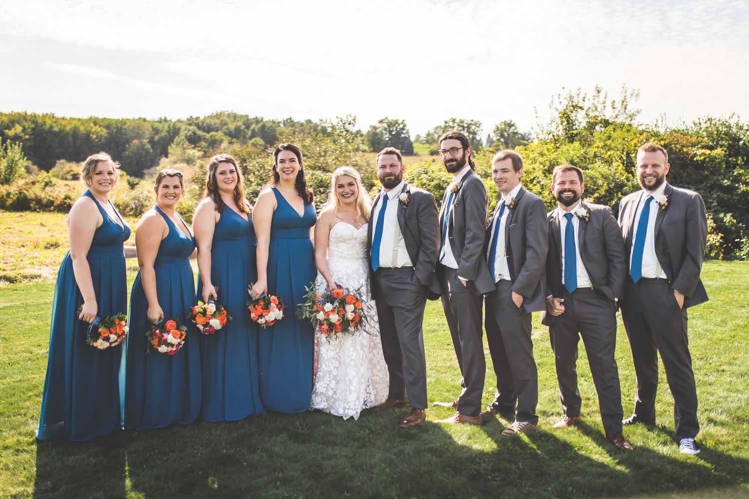 Wedding party lineup with bridesmaids in navy and groomsmen in gray suits on sunny field.