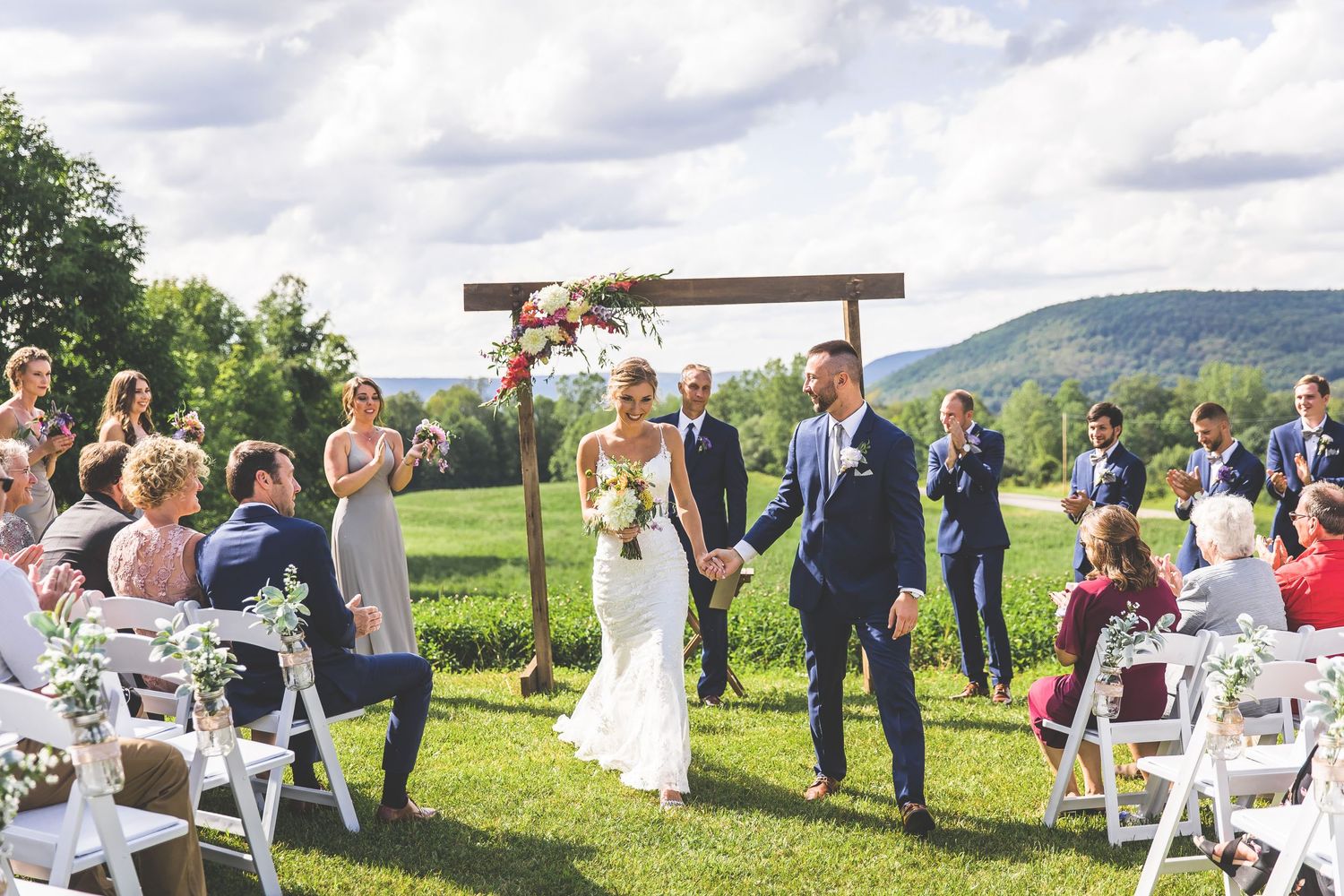 Panoramic view of outdoor wedding ceremony setup with wooden arch and white chairs on grassy hill.