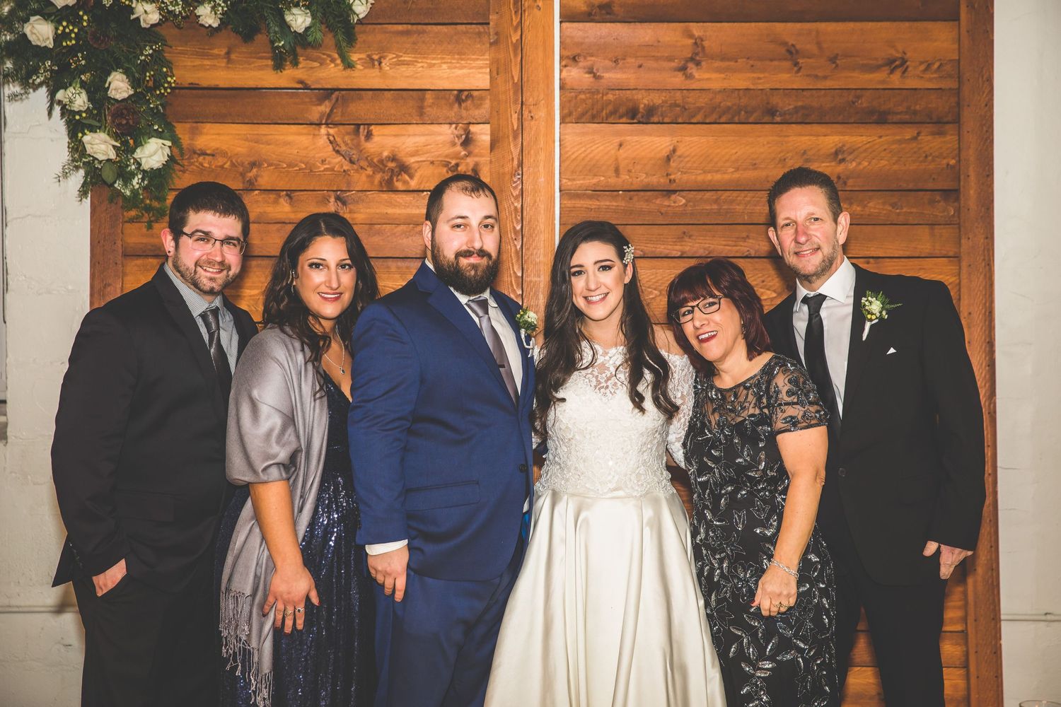 Group photo of wedding party members in formal attire against rustic wooden backdrop.