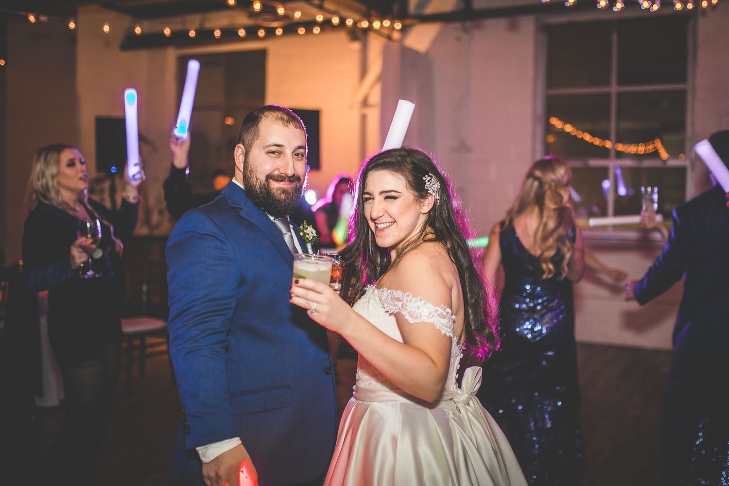 Wedding reception dance floor filled with guests dancing under string lights and colorful party lighting.