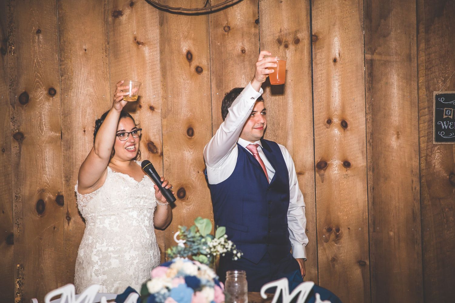 Couple cuts their wedding cake during rustic barn reception with string lights and wooden backdrop.